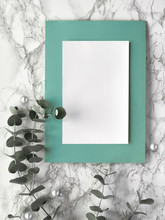 Christmas Frame With Fresh Eucalyptus Twigs And White Black Trinkets, Baubles With Stars. Flat Lay, Top View On Light Marble Background, Blank Paper Page With Text Space, Place For Your Text..