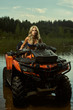 Beautiful blonde girl on a ATV rides on water and sand mountains. Green forest all around. In summer season