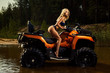 Beautiful blonde girl on a ATV rides on water and sand mountains. Green forest all around. In summer season