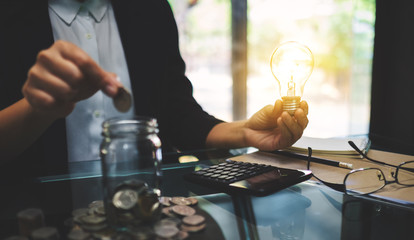 Wall Mural - Businesswoman holding a light bulb while putting coin into a glass jar for saving energy and money concept