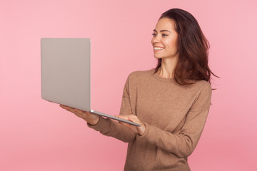 Wall Mural - Portrait of young woman in casual sweater standing, holding laptop and smiling while talking on video call, having online conference, freelancer working on computer. indoor studio shot isolated
