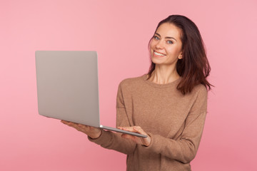 Wall Mural - Portrait of freelancer working on computer. Young woman in casual sweater holding laptop and smiling to camera, working or studying online using computer. studio shot isolated on pink background