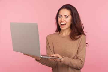Wall Mural - Portrait of young woman holding laptop and looking at camera with shocked expression, pleasantly surprised by computer software, shopping online. indoor studio shot isolated on pink background