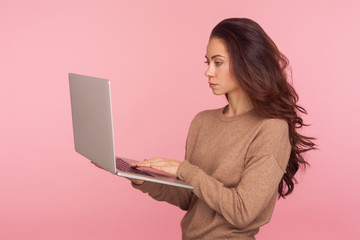 Wall Mural - Portrait of serious freelancer typing on laptop. Young woman in casual sweater working on computer with attentive focused expression, doing online business. studio shot isolated on pink background