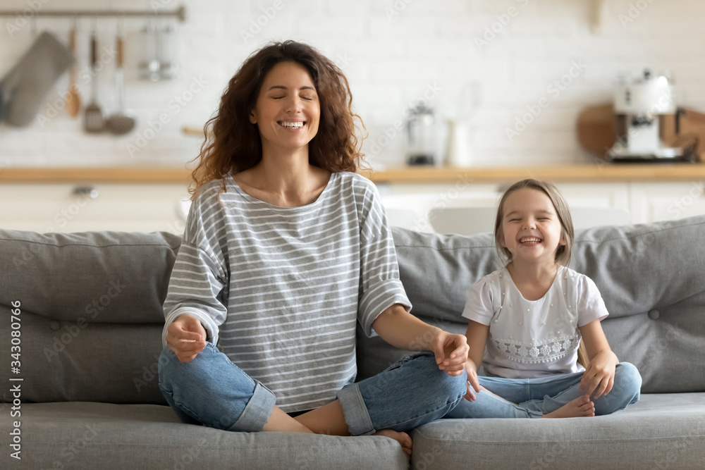 Full length happy young nanny mom sitting on sofa in lotus pose, teaching small daughter yoga breathing exercise. Friendly sincere two female generations family relaxing on couch, healthcare concept. - obrazy, fototapety, plakaty 