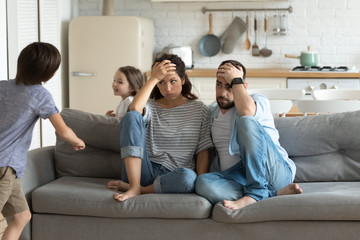 Stressed young parents sitting on couch, tired of crazy loud hyperactive little children siblings at home. Unhappy married couple irritated by naughty small kid son daughter, suffering from headache.