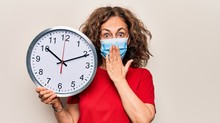 Middle Age Woman Wearing Coronavirus Protection Mask Holding Big Clock Ticking For Deadline Covering Mouth With Hand, Shocked And Afraid For Mistake. Surprised Expression