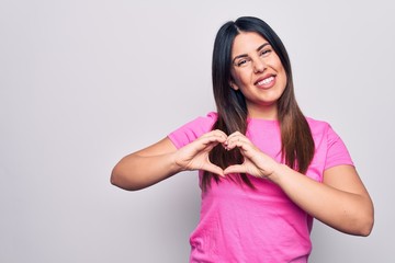 Poster - Young beautiful brunette woman wearing casual pink t-shirt standing over white background smiling in love doing heart symbol shape with hands. Romantic concept.