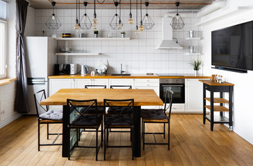 Modern scandinavian an eat-in kitchen interior design with big wooden table and chairs against light wood floor, bright white walls and furnitures with TV, appliances and hanging light bulbs