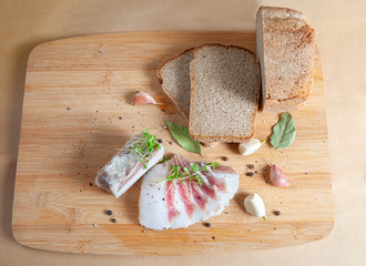 salted fat is cut into thin slices on top of sprouts of germinated mustard, next to it there is rye bread, garlic, pepper, bay leaf. On a wooden board