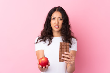 Wall Mural - Spanish Chinese woman over isolated pink background taking a chocolate tablet in one hand and an apple in the other