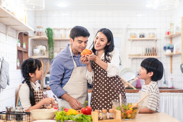 Wall Mural - Lovely cute Asian family making food in kitchen at home. Portrait of smiling mother, dad and children standing at cooking counter. Mom feeding dad some fruit with smile. Happy family activity together