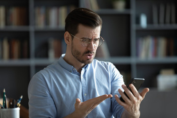 unhappy irritated young man wearing glasses having problem with broken or discharged smartphone, ind