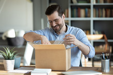 Smiling Man Wearing Glasses Unpacking Awaited Parcel, Looking Inside, Sitting At Work Desk, Satisfied Happy Customer Opening Cardboard Box With Online Store Order, Good Shipping Delivery Service