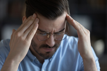 Close up unhealthy stressed young man wearing glasses touching forehead, suffering from headache, tired overworked employee, student massaging temples, feeling pain, health problem