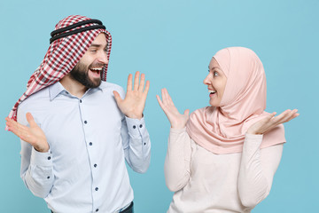 Wall Mural - Excited couple friends arabian muslim man wonam in keffiyeh kafiya ring igal agal hijab clothes isolated on blue background. People religious lifestyle concept. Looking at each other, spreading hands.