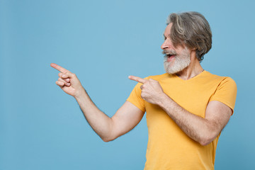 Excited elderly gray-haired mustache bearded man in casual yellow t-shirt posing isolated on blue background studio portrait. People lifestyle concept. Mock up copy space. Point index fingers aside.