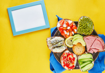 Poster - Assorted open-faced sandwiches, sandwiches with slices of sourdough with various fillings on a plate with a blue napkin and a yellow background top view. Flat lay. Frame for text - copy space