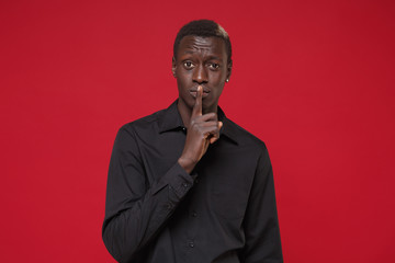 Secret young african american man guy in classic black shirt posing isolated on red background. People lifestyle concept. Mock up copy space. Saying hush be quiet with finger on lips shhh gesture.