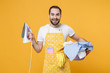 © ViDi Studio - Cheerful young man househusband in apron hold basket with clean clothes, iron doing housework isolated on yellow background studio portrait. Housekeeping concept. Mock up copy space. Looking camera.