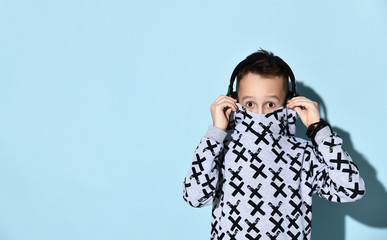 Wall Mural - Teenage kid in headphones and black bracelet. He is hiding his mouth in stylish hoodie while posing against blue background