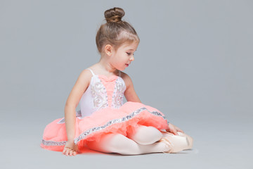 cute charming ballerina little girl in pink tutu is sitting on the floor trying to put on ballet sho