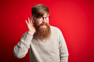 Canvas Print - Handsome Irish redhead man with beard wearing casual sweater and glasses over red background smiling with hand over ear listening an hearing to rumor or gossip. Deafness concept.