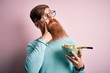 Redhead Irish healthy man with beard eating vegetarian green salad over pink background serious face thinking about question, very confused idea