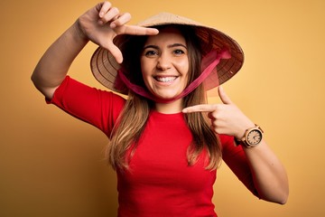 Canvas Print - Young blonde woman wearing traditional asian rice paddy straw hat over yellow background smiling making frame with hands and fingers with happy face. Creativity and photography concept.