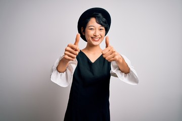 Young beautiful asian girl wearing casual dress and hat standing over isolated white background approving doing positive gesture with hand, thumbs up smiling and happy for success. Winner gesture.