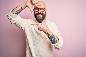 Sticker - Handsome bald man with beard and tattoo wearing glasses and sweater over pink background smiling making frame with hands and fingers with happy face. Creativity and photography concept.