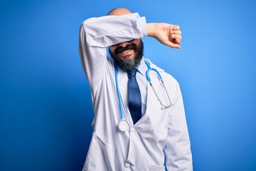 Canvas Print - Handsome bald doctor man with beard wearing glasses and stethoscope over blue background covering eyes with arm smiling cheerful and funny. Blind concept.