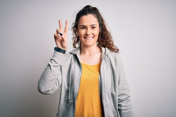 Wall Mural - Young beautiful sportswoman with curly hair wearing sportswear over white background smiling with happy face winking at the camera doing victory sign. Number two.