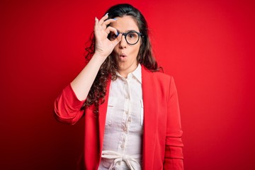 Sticker - Young beautiful woman with curly hair wearing jacket and glasses over red background doing ok gesture shocked with surprised face, eye looking through fingers. Unbelieving expression.