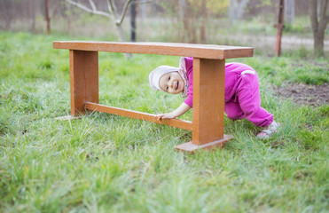 Wall Mural - Happy little child girl plays peekaboo under wooden bench in garden