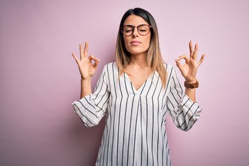 Poster - Young beautiful woman wearing casual striped t-shirt and glasses over pink background relaxed and smiling with eyes closed doing meditation gesture with fingers. Yoga concept.