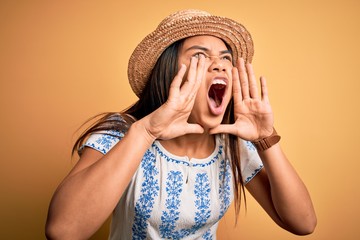 Young beautiful asian girl wearing casual t-shirt and hat standing over yellow background Shouting angry out loud with hands over mouth