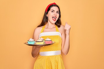 Poster - Young beautiful pin up woman wearing 50s fashion vintage dress holding tray with cupcakes screaming proud and celebrating victory and success very excited, cheering emotion