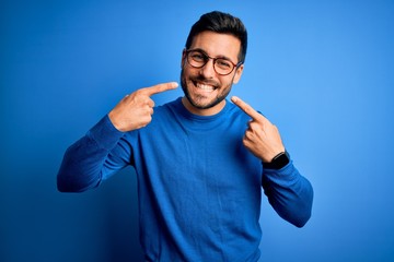 Poster - Young handsome man with beard wearing casual sweater and glasses over blue background smiling cheerful showing and pointing with fingers teeth and mouth. Dental health concept.