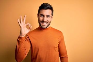 Young handsome man with beard wearing casual sweater standing over yellow background smiling positive doing ok sign with hand and fingers. Successful expression.