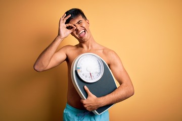 Poster - Handsome man shirtless wearing swimwear holding weight machine over yellow background with happy face smiling doing ok sign with hand on eye looking through fingers