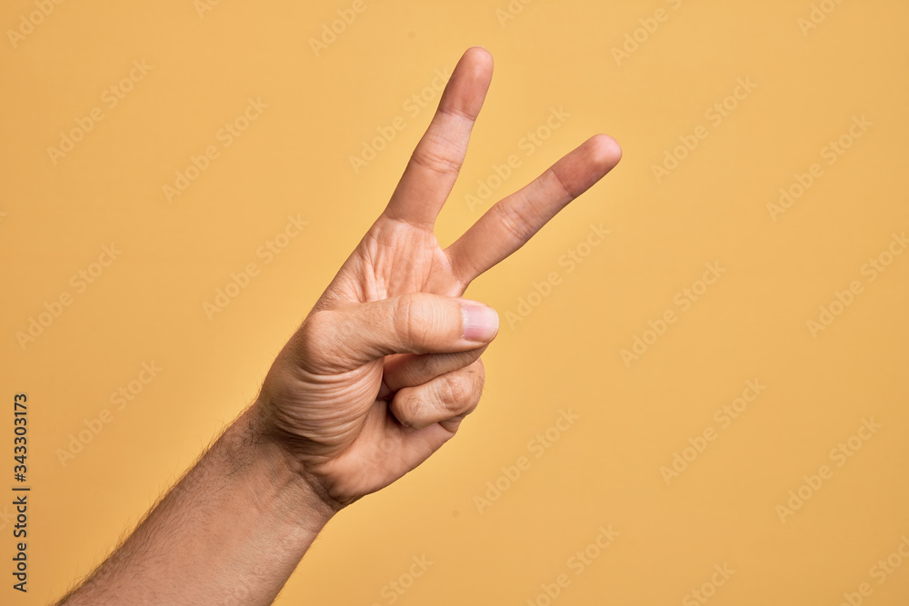 Hand of caucasian young man showing fingers over isolated yellow background counting number 2 showing two fingers, gesturing victory and winner symbol - obrazy, fototapety, plakaty 