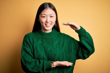 Sticker - Young beautiful asian woman wearing green winter sweater over yellow isolated background gesturing with hands showing big and large size sign, measure symbol. Smiling looking at the camera. Measuring