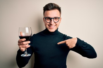 Poster - Young handsome caucasian man drinking an alcoholic glass of red wine over isolated background with surprise face pointing finger to himself