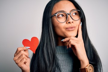 Wall Mural - Young beautiful chinese woman holding paper heart over isolated white background serious face thinking about question, very confused idea