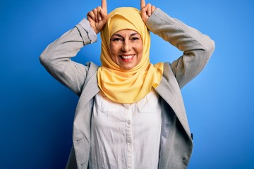 Wall Mural - Middle age brunette business woman wearing muslim traditional hijab over blue background doing funny gesture with finger over head as bull horns