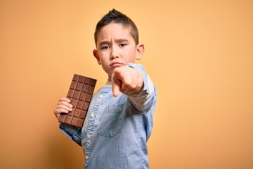 Wall Mural - Young little boy kid eating sweet chocolate bar for dessert over isolated yellow background pointing with finger to the camera and to you, hand sign, positive and confident gesture from the front