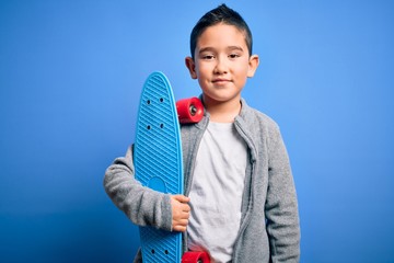 Poster - Young little boy kid skateboarder holding modern skateboard over blue isolated background with a happy face standing and smiling with a confident smile showing teeth