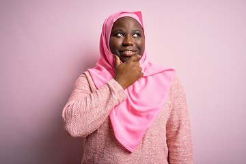 Poster - Young african american plus size woman wearing muslim hijab over isolated pink background with hand on chin thinking about question, pensive expression. Smiling with thoughtful face. Doubt concept.