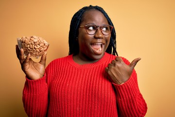 Sticker - Young african american plus size woman with braids holding bowl with healthy peanuts pointing and showing with thumb up to the side with happy face smiling